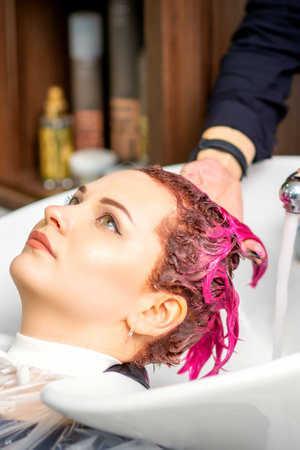 Washing dyed female hair. A young caucasian woman having her hair washed in a beauty salon. Professional hairdresser washes pink color paint off of a customers hairの写真素材