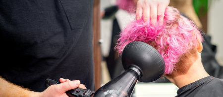 Drying short pink bob hairstyle of a young caucasian woman with a black hair dryer with the brush by hands of a male hairdresser in a hair salon, close upの写真素材