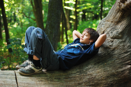 Teenage boy is lying on trunk and listening to music  Gadget is on his chest  Small earphones is in his ears  Green forest is full of sunlight and blurred の写真素材