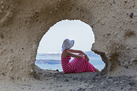 Woman in a long dress sitting on  rocks of  cliff, near  cave in the shape of a heart. Santorini, Greeceの写真素材