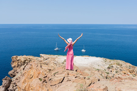 Woman in a long summer dress standing on a cliff and watches the ships at sea. Greece, Santorini.