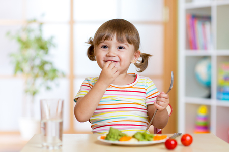 Happy child eats vegetables sitting at table in nursery Happy child eats vegetables sitting at table in nursery