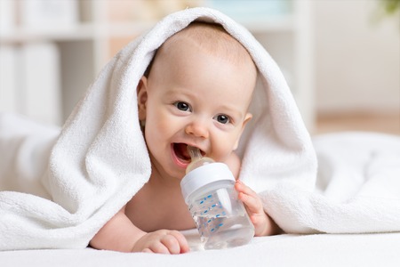 Happy baby boy drinks water from bottle wrapped towel after bath