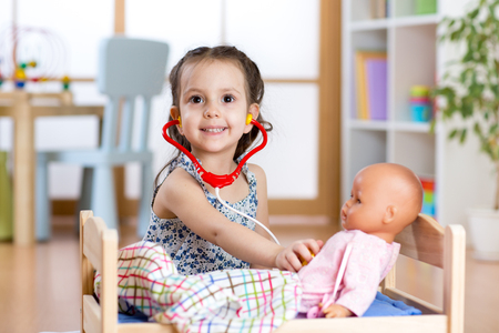 child girl playing doctor role game examining a doll using stethoscope sitting in playroom at home, school or kindergartenの写真素材