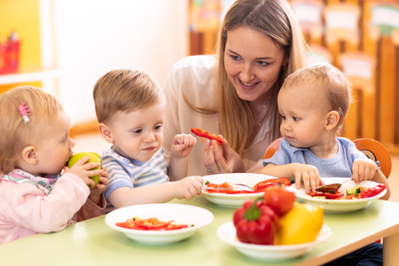 Kids have a lunch in daycare centre. Children eating healthy food in kindergarten. Nursery teacher with babies at tableの写真素材
