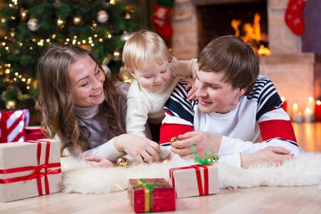 Happy young family at christmas eve. Mom, dad and baby lying down with gift boxes near xmas tree and fireplaceの写真素材