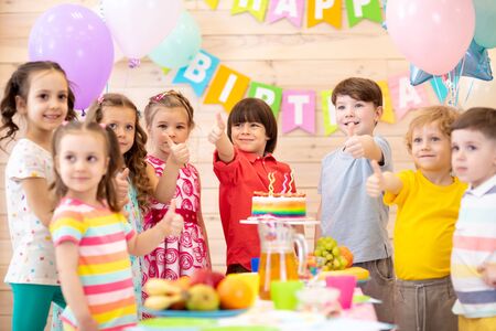 Preschool children celebrating birthday party. Kids stand arround festive table and show thumbs upの写真素材