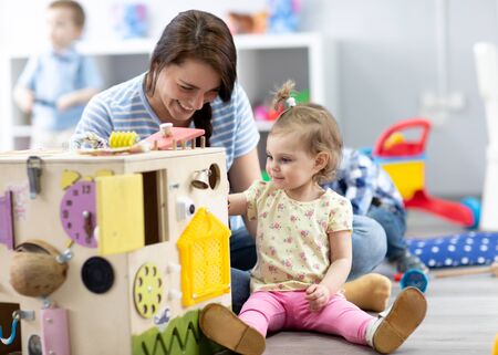 Cute toddler baby girl playing with busyboard. Teacher or babysitter teaching kid in nursery. Children's educational toys.の写真素材