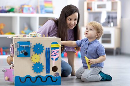 Cute toddler baby and mom playing with busy board at home or daycare. Childrens educational toys. Wooden game diy boardの写真素材