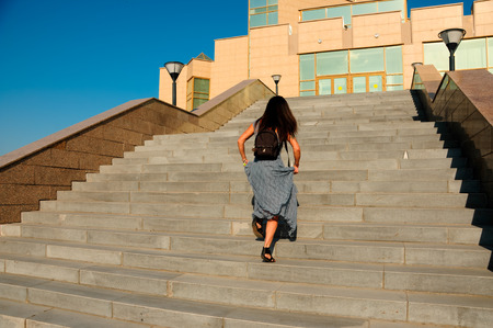 brown-haired woman with long hair and a long gray dress on a city street climbs the stairs in the...
