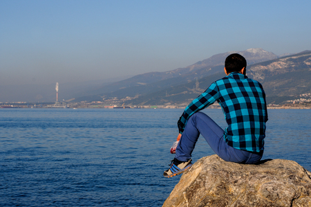 Turkey January 22, 2017: young men in a shirt in a cage sits on a stone near the coast of the blue sea against a background of  the mountains on a sunny afternoon Iskenderunのeditorial素材