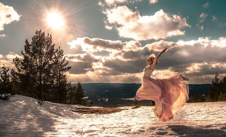 Blonde girl in a pink long dress in a jump on top of a mountain against the sky and clouds and sunの写真素材
