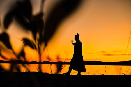 Silhouette of a girl in a dress with a phone in his hand against the backdrop of a beautiful orange sunset on the shore of Lake Zyuratkul, Chelyabinsk Region, Russiaの写真素材