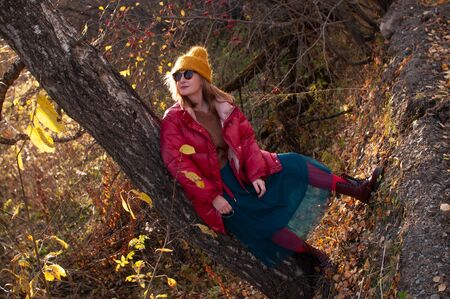 Blonde girl in a bright red jacket and a yellow hat in the autumn forest at sunsetの写真素材