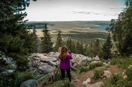 tourist woman stot on top of a mountain in a coniferous forest and looks at the landscape in summer, view from the back Russia, Urals, Zyuratkul Ridgeの写真素材