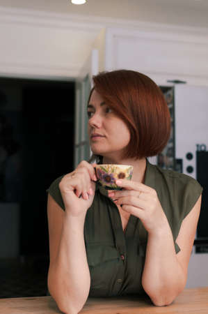 Portrait of a woman with bob haircut and red natural hair in everyday life at home with a cup of tea in her handsの写真素材