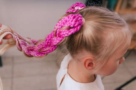 Girl with blond hair of five years old with a hairstyle of pink braids indoors, view from the backの写真素材