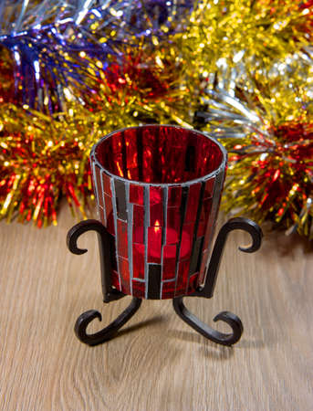 Candlestick from red glass on a metal stand. Candlestick stands on a wooden table. Behind the background of the Christmas tree tinsel.の写真素材