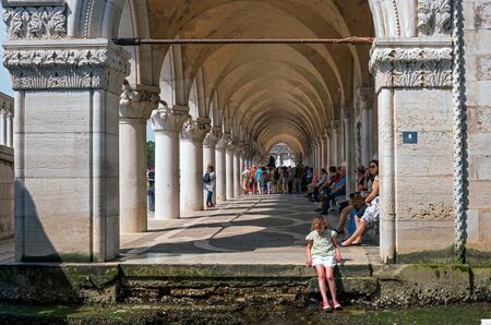 Venice, Italy - 08 May 2018: Arcade of Palazzo Ducale at Piazza San Marco. The palace was the residence of the Doge of Venice. Tourists in line for tickets. Girl sitting on the edge of the channelのeditorial素材