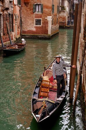 Venice, Italy - October 13, 2017: Gondolier stopped in anticipation of passengers. Gondolier in a striped sweater and a straw hat. The gondola is richly decorated with red carpets.のeditorial素材