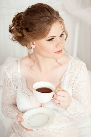 Girl holding a white coffee mug. In a white coat. Coffee in bed. Morning begins with coffee. Coffee in the hands of the brideの写真素材
