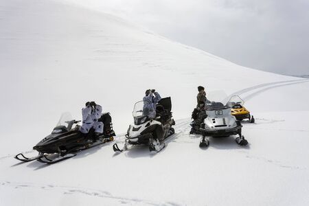 Hunters in winter camouflage on snowmobiles are looking through binoculars. Four hunters look through the binoculars on the mountainside in search of a hunting object. Kamchatka, Siberia, Russiaの写真素材