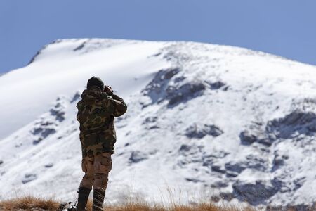 Hunter in the mountains in camouflage looks through binoculars. A man in a hunting camouflage jacket looks at the mountainside in search of animals.の写真素材