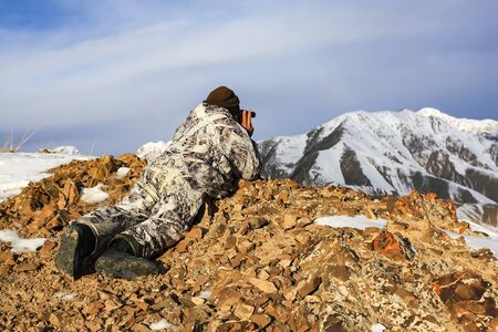 Hunter in winter camouflage in the mountains lying watching with binoculars. A huntsman lies in the mountains in winter and uses an optical device to view hunting animals.の写真素材