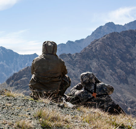 Two men in camouflage with weapons are watching in the mountains. Hunters on the mountain slope secretly search for hunting objects.の写真素材