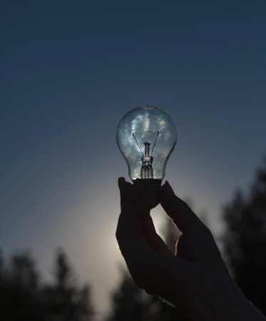 Incandescent lamp of traditional shape in woman's hand against the sky. An electric light bulb is typical source of artificial light.の写真素材