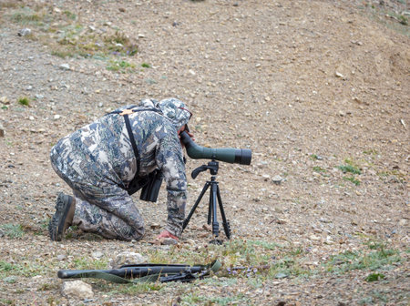 Hunter in camouflage looks carefully through telescope on tripod in mountainous area. Ranger secretly observes the situation in the mountains through an optical device.の写真素材