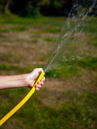 Yellow watering hose with jet of water in the hand of teenager watering the lawn. Typical hosing in hot summer weather in country garden.の写真素材