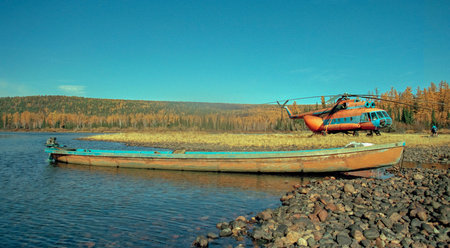 Homemade boat with an outboard motor and Soviet helicopter in upper reaches of Siberian river in autumn. Long boat with an old engine on the rocky shore of shallow river by sitting helicopter.の写真素材