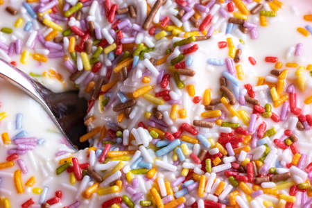 Close-up of a mug with yoghurt and colored sprinkles made of sugar, a spoon in the white cream,...