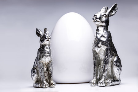two silver mate easter bunny figures in front of a white ceramic egg, creative easter and spring...