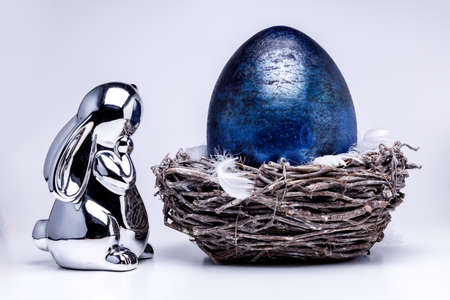 Silver metalic easter bunny figure looks into a royal blue easter egg laying in a easter nest made of brown branches, filled with white feahters on white uniform backgroundの写真素材