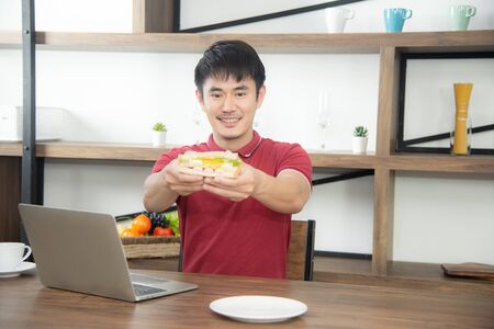 The business man with casual  red t-shirt having breakfast and eating sandwich, drinking coffee. Young man working on labtop or computer  in the loft style kitchen roomの写真素材