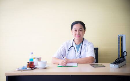The closed up woman doctor with white uniform  and medical devices such as stethoscope and blood pressure device is checking up the patient health and taking note on the white paper at the hospital or clinic wihic is the healthcare businessの写真素材