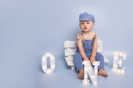 One year birthday party decorations. Little boy in a blue cap on the white bench on the background of a blue wall. Happy child playing with glowing letters one. blue background.の写真素材