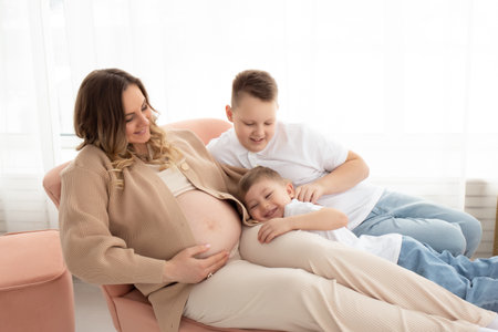a cute little boy hugs his mother for a pregnant belly on a sofa in a large bright living room....