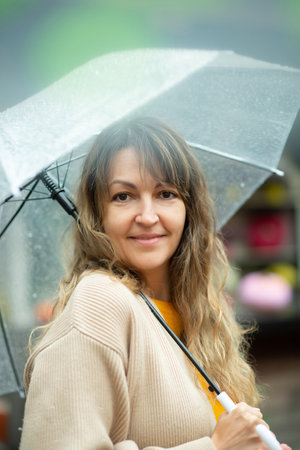 close-up portrait of a girl looking into the frame in a beige knitted sweater with a transparent umbrella in her hands. girl with umbrella smiling in the rain.の写真素材