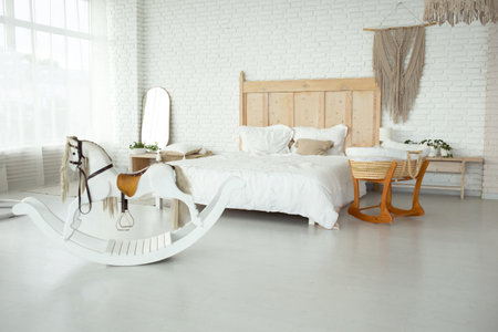 large bright bedroom with white walls and a large bed with a white knitted bedspread. near the bed there is a small wicker cradle with a toy and a beautiful toy rocking horse. family expecting pregnancy.の写真素材