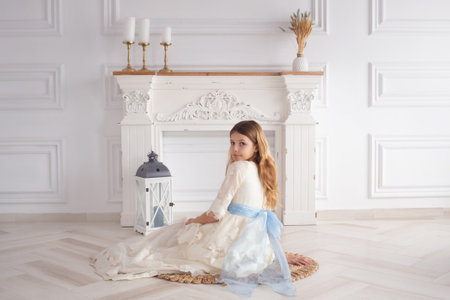 A beautiful girl in a smart white dress sits on the floor near the fireplace with candles on the day of her first communion.の写真素材