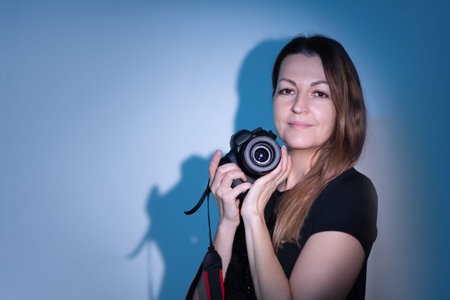 A woman stands holding a camera in her hands. She is smiling slightly while posing against a blue...