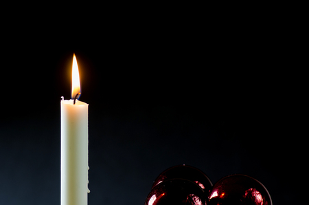 A white burning candle with red christmas balls at a black backgroundの写真素材