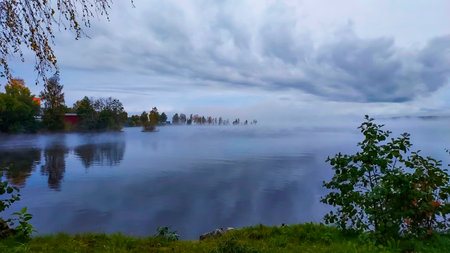 Beautiful autumn landscape with lake and trees in the fog on a cloudy dayの写真素材
