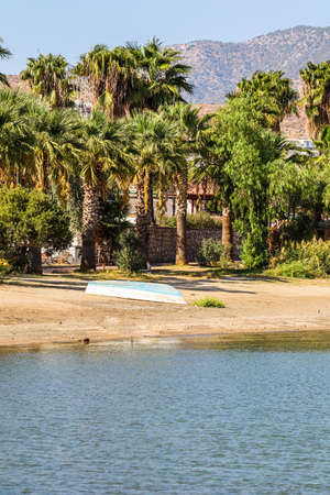 Blue sea, boat on the sandy beach, houses, palms and mountains on the Aegean coast. Summer holidays and coastal nature conceptの写真素材