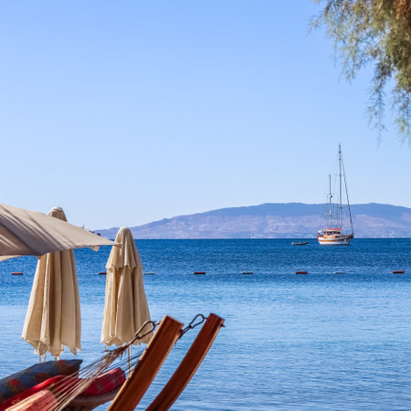 Beach umbrellas and sun loungers on the shore of a calm blue sea bay with a sailboat. Summer vacation and recreation area conceptの写真素材