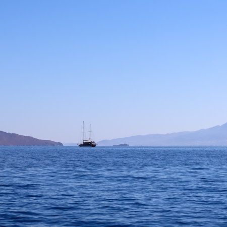 A calm blue sea with a traditional sailing boat in the distance, against a backdrop of hazy mountains and a clear sky. High quality photoの写真素材