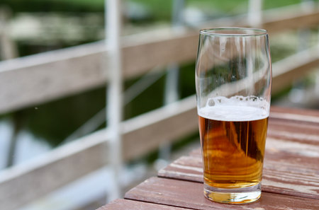 Pint glass partially filled with golden lager beer and white foam on a rustic wooden table, enjoying...